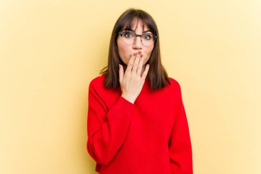 Sarı arka planda izole edilmiş genç beyaz kadın şok olmuş, ağzı ellerle kaplanmış, yeni bir şey keşfetmeye hevesli..