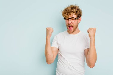 Mavi arka planda izole edilmiş genç beyaz bir adam zaferden sonra yumruğunu kaldırıyor, kazanan konsept..