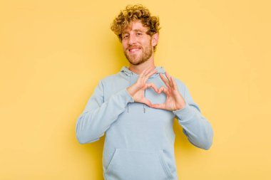 Sarı arka planda izole edilmiş genç beyaz adam gülümsüyor ve elleriyle kalp şeklini gösteriyor..