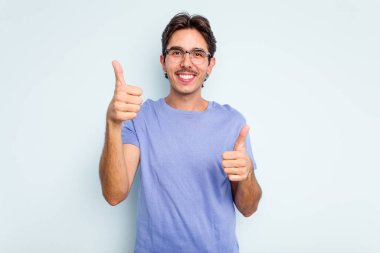 Genç İspanyol adam mavi arka planda izole edilmiş. Baş parmağını kaldırıyor, gülümsüyor ve kendinden emin..