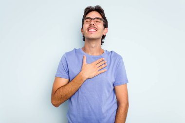 Mavi arka planda izole edilmiş genç İspanyol adam yüksek sesle gülüyor ve elini göğsüne koyuyor..