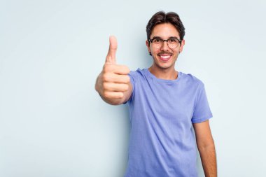 Genç İspanyol adam mavi arka planda izole edilmiş gülümsüyor ve başparmağını kaldırıyor.