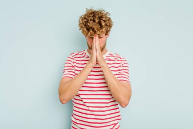 Kafkasyalı genç adam mavi arka planda izole edilmiş dua ediyor, bağlılık gösteriyor, dindar biri ilahi ilham arıyor..