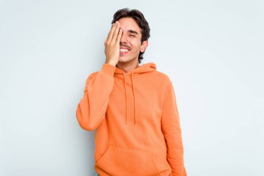 Genç İspanyol adam mavi arka planda izole edilmiş. Yüzünün yarısını avuç içiyle kaplayarak eğleniyor..