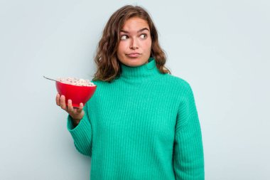 Mavi arka planda izole edilmiş bir kase mısır gevreği tutan genç beyaz kadın şaşkın ve kararsız hissediyor..