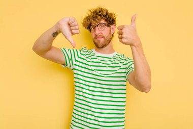 Sarı arka planda izole edilmiş genç beyaz adam baş parmağını kaldırıyor ve baş parmak aşağı, zor seçim konsepti.