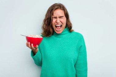 Mavi arka planda izole edilmiş bir kase mısır gevreği tutan genç beyaz kadın çok sinirli ve agresif bağırıyor..