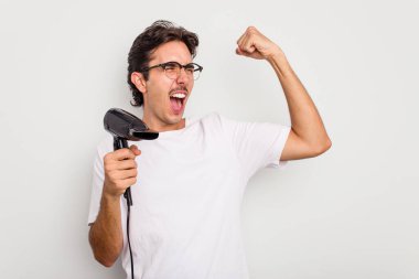 Beyaz arka planda izole edilmiş saç kurutma makinesi tutan İspanyol genç adam zafer kazandıktan sonra yumruğunu kaldırdı..
