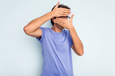 Mavi arka planda izole edilmiş genç İspanyol adam kameraya parmaklarıyla göz kırpıyor, yüzünü kapatmaktan utanıyor..