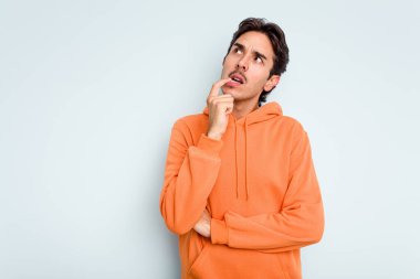 Genç İspanyol adam mavi arka planda izole edilmiş şüpheli ve şüpheci bir ifadeyle yana bakıyor..