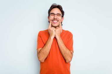 Mavi arka planda izole edilmiş genç İspanyol adam gülümsüyor, parmaklarını ağıza doğrultuyor..