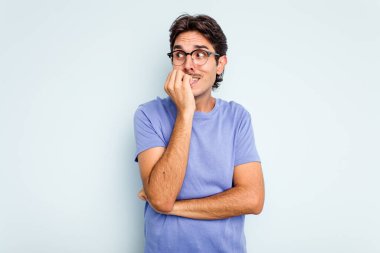 Mavi arka planda izole edilmiş, tırnak yiyen, gergin ve endişeli genç bir adam..