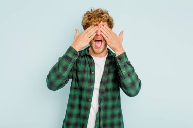 Mavi arka planda soyutlanmış genç beyaz bir adam elleriyle gözlerini kapatıyor, geniş bir gülümsemeyle bir sürpriz bekliyor..