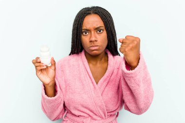 Bornoz giyen genç Afrikalı bir kadın mavi arka planda izole edilmiş bir deodorant tutuyor. Kameraya yumruk, agresif yüz ifadesi gösteriyor..