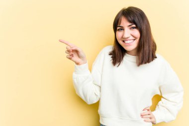 Sarı arka planda izole edilmiş genç beyaz kadın işaret parmağıyla işaret ederek gülümsüyor..