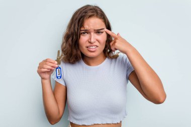 Young caucasian woman holding home keys isolated on blue background showing a disappointment gesture with forefinger.