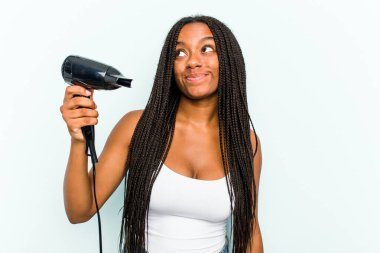 Genç Afrikalı Amerikalı bir kadın elinde saç kurutma makinesiyle mavi arka planda izole bir şekilde hedeflerine ve amaçlarına ulaşma hayali kuruyor.