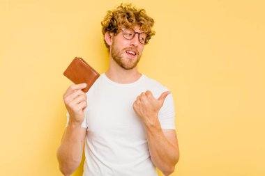 Sarı arka plan noktalarında cüzdan tutan genç beyaz bir adam başparmak uzakta, gülüyor ve kaygısız..