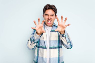 Mavi arka planda izole edilmiş genç İspanyol bir adam pençelerini bir kediyi taklit ederken gösteriyor, agresif bir hareket..