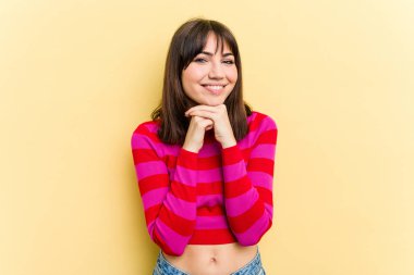 Sarı arka planda izole edilmiş genç beyaz kadın ellerini çenesinin altında tutuyor, mutlu bir şekilde yana bakıyor..