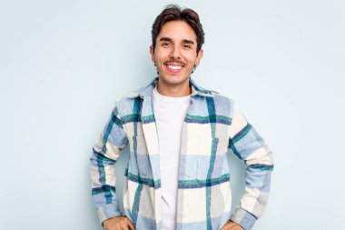 Latin kökenli genç adam mavi arka planda izole edilmiş. Kendine güveniyor. Kalçaları tutuyor..