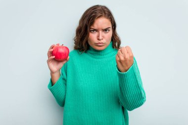 Mavi arka planda izole edilmiş, yumruğunu kameraya gösteren beyaz bir kadın, agresif bir yüz ifadesi..