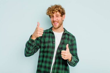 Kafkasyalı genç adam mavi arka planda izole edilmiş. Baş parmağını kaldırıyor, gülümsüyor ve kendinden emin..