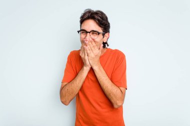 Genç İspanyol bir adam mavi arka planda izole edilmiş bir şekilde bir şeye gülüyor, elleriyle ağzını kapatıyor..