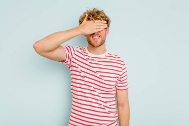 Mavi arka planda soyutlanmış genç beyaz bir adam elleriyle gözlerini kapatıyor, geniş bir gülümsemeyle bir sürpriz bekliyor..