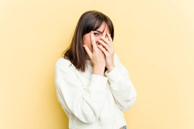 Sarı arka planda izole edilmiş genç beyaz kadın parmaklarının arasından korkmuş ve gergin bir şekilde göz kırp..