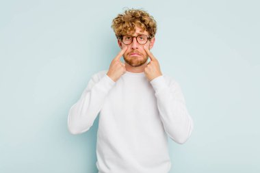 Kafkasyalı genç adam mavi arka planda izole edilmiş, ağlıyor, bir şeyden memnun değil, acı ve kafa karışıklığı konsepti..