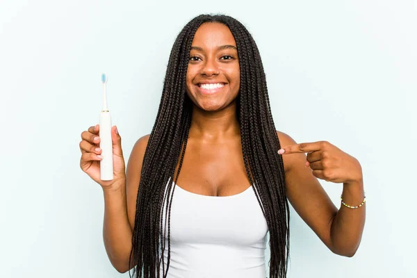 年轻的非洲裔美国女人拿着电动牙刷 与蓝色背景的人隔离 手指着衬衫的复制品空间 自豪而自信 — 图库照片