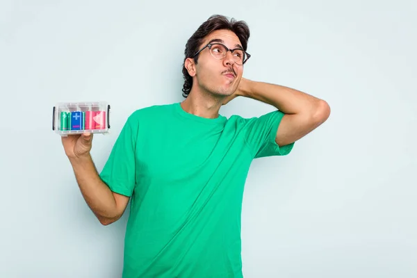 Young hispanic man holding battery box isolated on white background touching back of head, thinking and making a choice.