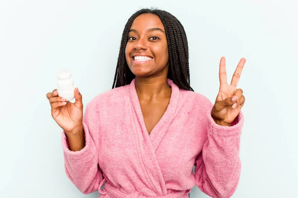 身穿浴衣的年轻非洲裔美国妇女拿着一种除臭剂 用蓝色背景隔离 快乐而无忧无虑 用手指展示和平的象征 — 图库照片