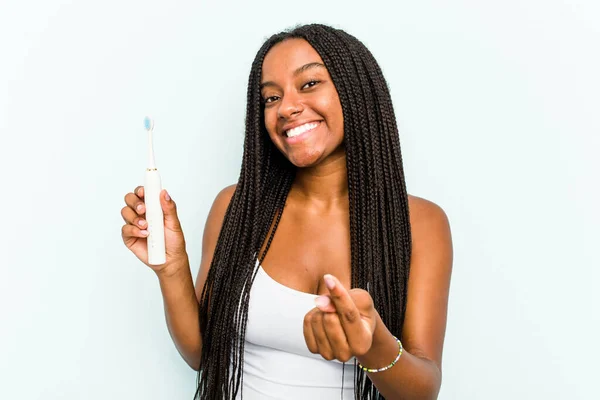 年轻的非洲裔美国女人拿着用蓝色背景隔开的电动牙刷 用手指指著你 好像邀请你走近些 — 图库照片