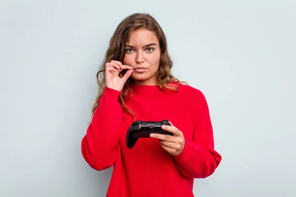 Young caucasian gamer woman holding a game controller isolated on blue background with fingers on lips keeping a secret.