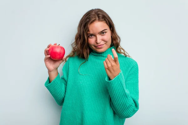 年轻的高加索女人 有一个蓝色背景的苹果 用手指指着你 好像邀请你走近些 — 图库照片