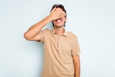 Mavi arka planda izole edilmiş genç İspanyol adam elleriyle gözlerini kapatıyor, geniş bir gülümsemeyle bir sürpriz bekliyor..