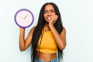 Genç Afrikalı Amerikalı bir kadın mavi arka planda izole edilmiş bir saat tutuyor tırnaklarını yiyor, gergin ve endişeli..