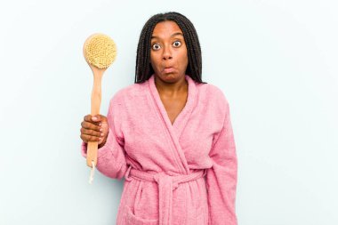 Elinde küvet fırçası tutan genç Afrikalı bir kadın mavi arka planda izole edilmiş omuzlarını silkeliyor ve gözleri açık..