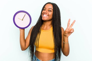 Genç Afrikalı Amerikalı bir kadın mavi arka planda izole edilmiş bir saat tutuyor neşeli ve kaygısız parmakları olan bir barış sembolü gösteriyor..
