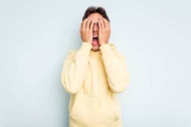 Genç İspanyol adam mavi arka planda izole edilmiş. Yüzünün yarısını avuç içiyle kaplayarak eğleniyor..