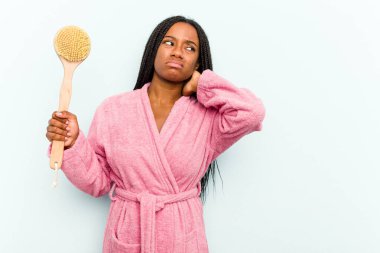 Genç Afrikalı Amerikalı bir kadın elinde bir küvet fırçası tutuyor mavi arka planda izole edilmiş, kafasının arkasına dokunuyor, düşünüyor ve bir seçim yapıyor..