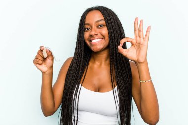 Elinde işitme cihazı tutan genç Afro-Amerikalı kadın mavi arka planda izole edilmiş neşeli ve kendinden emin..