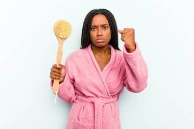 Elinde küvet fırçası olan genç Afrikalı bir kadın mavi arka planda izole edilmiş. Kameraya yumruk, agresif yüz ifadesi gösteriyor..