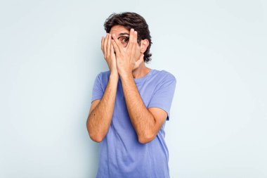 Genç İspanyol adam mavi arka planda izole edilmiş. Korkmuş ve gergin parmaklarıyla göz kırp..