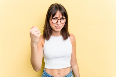 Sarı arka planda izole edilmiş genç beyaz kadın kameraya yumruğunu gösteriyor, agresif yüz ifadesi..