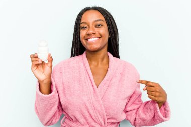 Üzerinde deodorant olan genç Afrikalı bir kadın mavi arka plandaki izole edilmiş bir deodorant tutuyor. Bir gömlek fotokopi alanını işaret ediyor, gururlu ve kendinden emin.