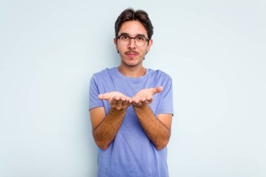 Genç İspanyol bir adam mavi arka planda izole edilmiş. Elinde avuç içi olan bir şeyle kameraya sunuyor..