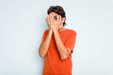Genç İspanyol adam mavi arka planda izole edilmiş. Korkmuş ve gergin parmaklarıyla göz kırp..
